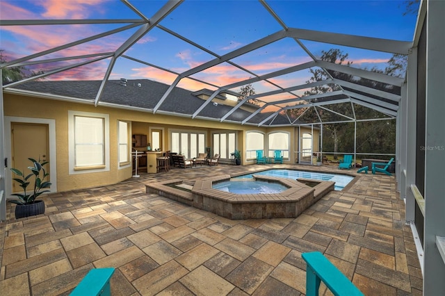 pool at dusk featuring glass enclosure, an in ground hot tub, and a patio area