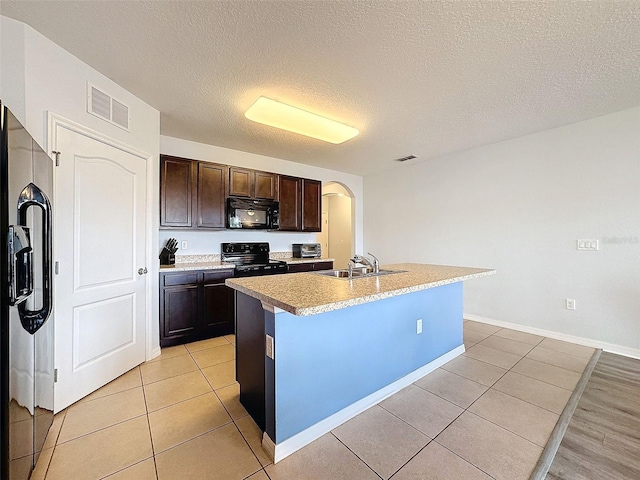 kitchen with a kitchen island with sink, black appliances, sink, dark brown cabinets, and light tile patterned flooring