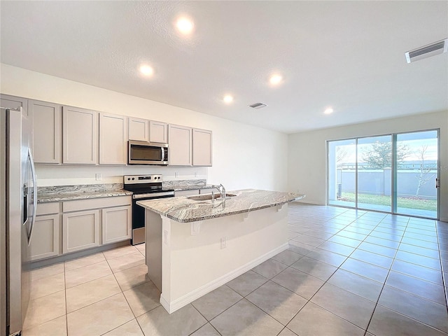 kitchen with gray cabinets, an island with sink, appliances with stainless steel finishes, light tile patterned flooring, and light stone countertops