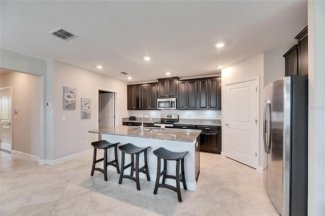 kitchen featuring light stone counters, an island with sink, decorative backsplash, appliances with stainless steel finishes, and sink