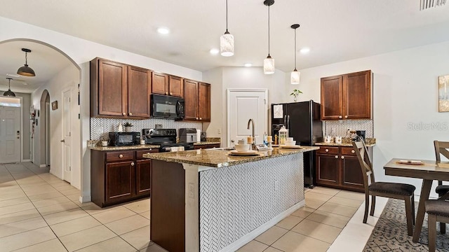 kitchen featuring stone counters, light tile patterned floors, black appliances, hanging light fixtures, and a kitchen island with sink