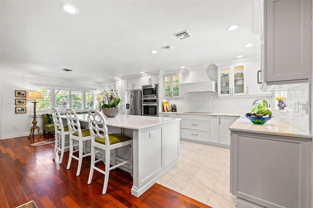 kitchen featuring custom exhaust hood, appliances with stainless steel finishes, glass insert cabinets, white cabinets, and a kitchen island