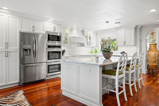 kitchen with arched walkways, stainless steel appliances, glass insert cabinets, white cabinets, and premium range hood