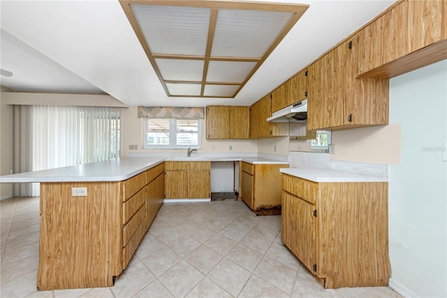 kitchen featuring light tile patterned flooring, under cabinet range hood, a peninsula, light countertops, and brown cabinetry
