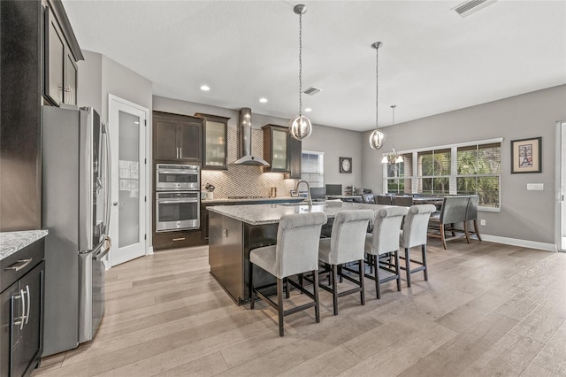 kitchen with appliances with stainless steel finishes, a kitchen bar, a kitchen island with sink, dark brown cabinets, and wall chimney range hood