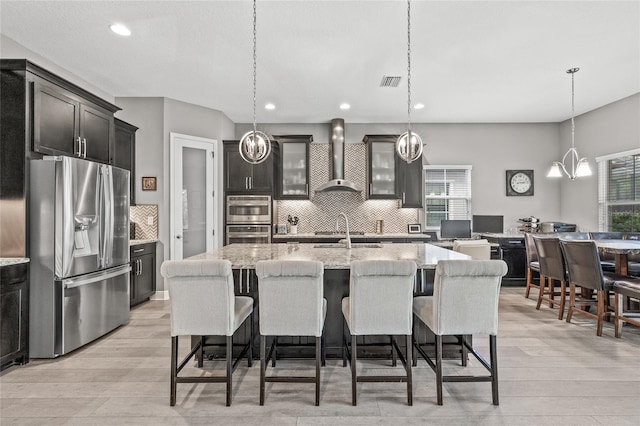 kitchen featuring wall chimney exhaust hood, stainless steel appliances, hanging light fixtures, and a center island with sink