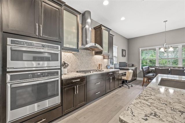 kitchen with wall chimney exhaust hood, dark brown cabinetry, appliances with stainless steel finishes, light hardwood / wood-style floors, and backsplash