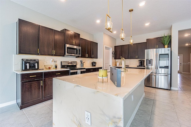 kitchen with stainless steel appliances, a kitchen island with sink, decorative light fixtures, light tile patterned flooring, and light stone counters