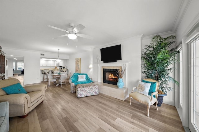 living room with crown molding, a tile fireplace, ceiling fan, a healthy amount of sunlight, and light wood-type flooring