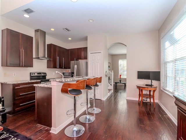 kitchen with appliances with stainless steel finishes, dark hardwood / wood-style floors, light stone counters, a center island with sink, and wall chimney exhaust hood