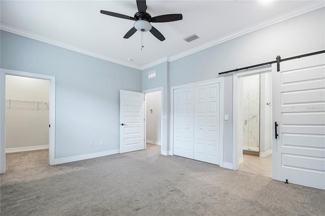 unfurnished bedroom with light carpet, ceiling fan, a barn door, a spacious closet, and ornamental molding