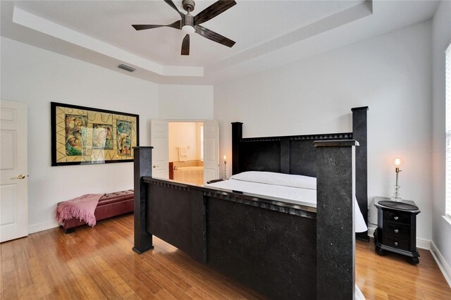 bedroom with wood-type flooring, ensuite bathroom, ceiling fan, and a tray ceiling