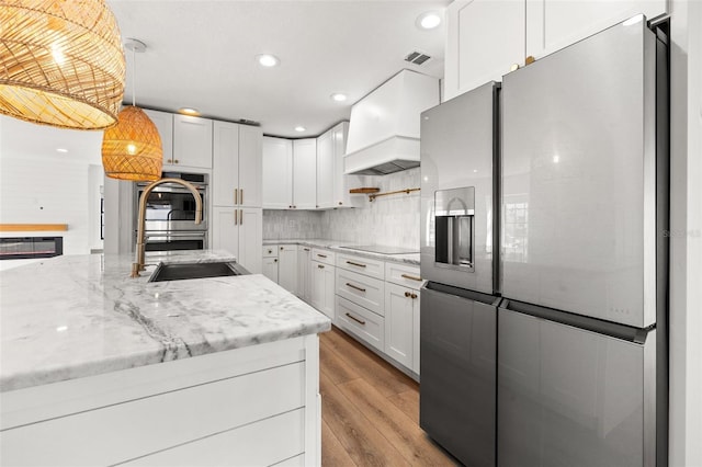 kitchen with custom exhaust hood, white cabinetry, decorative light fixtures, light wood-type flooring, and appliances with stainless steel finishes