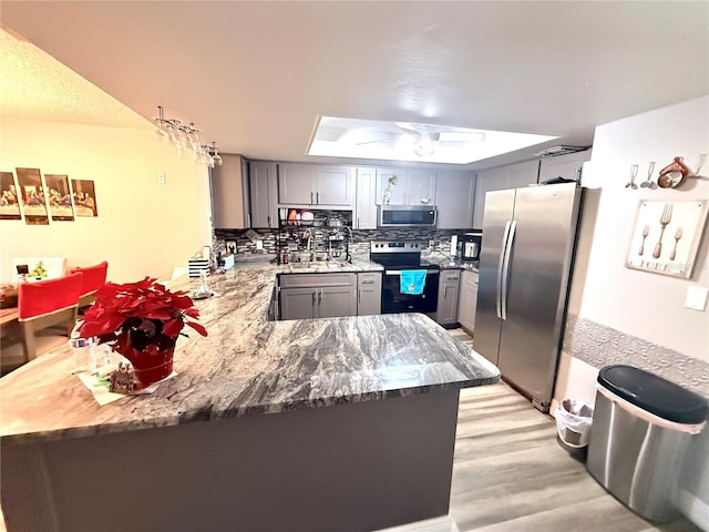 kitchen with appliances with stainless steel finishes, backsplash, kitchen peninsula, a raised ceiling, and gray cabinetry