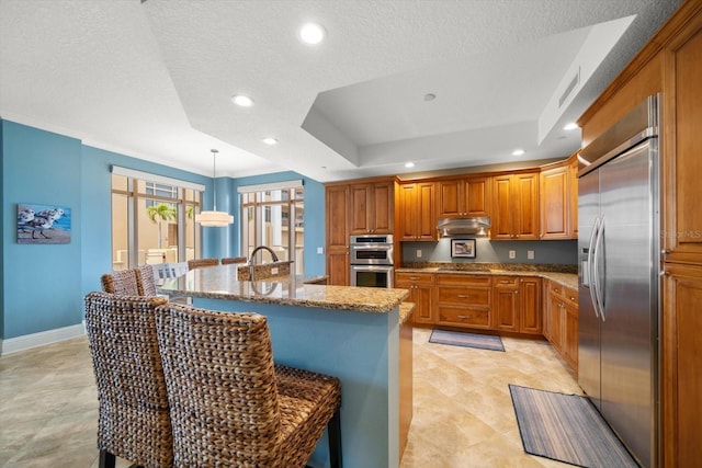 kitchen featuring a kitchen bar, an island with sink, stainless steel appliances, a tray ceiling, and hanging light fixtures