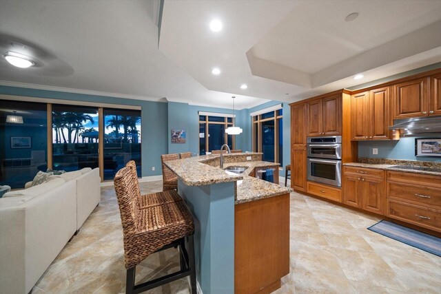 kitchen featuring a breakfast bar, hanging light fixtures, a kitchen island with sink, light stone countertops, and stainless steel double oven