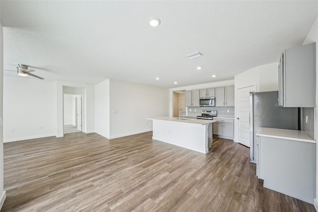 kitchen with stainless steel appliances, an island with sink, decorative backsplash, gray cabinets, and hardwood / wood-style flooring