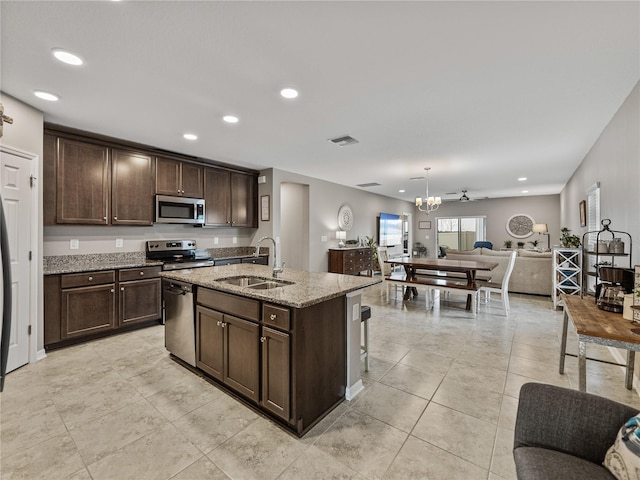 kitchen with appliances with stainless steel finishes, a kitchen island with sink, light stone counters, dark brown cabinetry, and sink