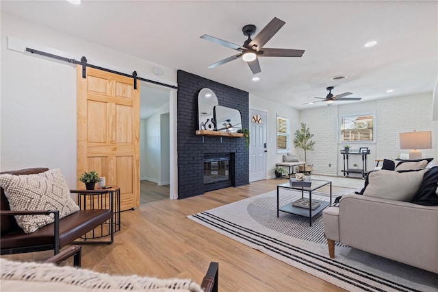 living room with ceiling fan, a fireplace, brick wall, a barn door, and light wood-type flooring