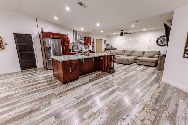 kitchen featuring lofted ceiling, light stone counters, a center island with sink, stainless steel fridge, and wall chimney range hood