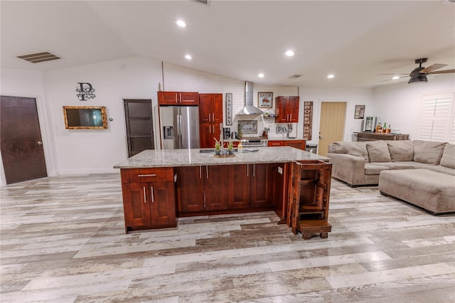 kitchen with wall chimney range hood, appliances with stainless steel finishes, light stone counters, a center island with sink, and vaulted ceiling