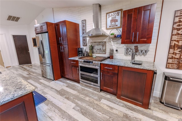 kitchen with wall chimney range hood, stainless steel appliances, light stone countertops, decorative backsplash, and light wood-type flooring