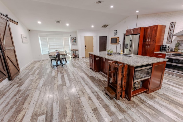 kitchen with an island with sink, sink, light stone countertops, stainless steel appliances, and a barn door