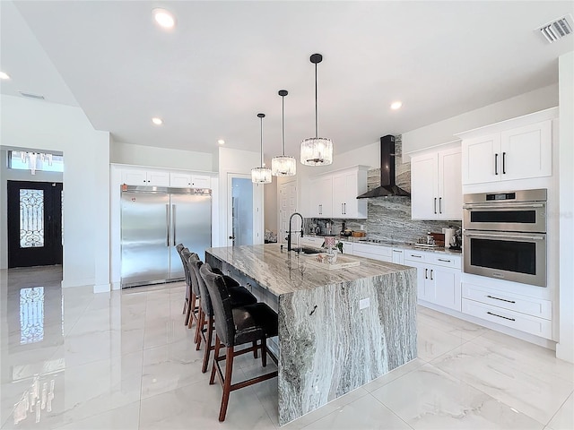 kitchen featuring white cabinets, appliances with stainless steel finishes, wall chimney exhaust hood, sink, and a kitchen island with sink