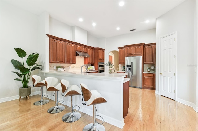 kitchen featuring arched walkways, stainless steel appliances, light countertops, under cabinet range hood, and a kitchen bar