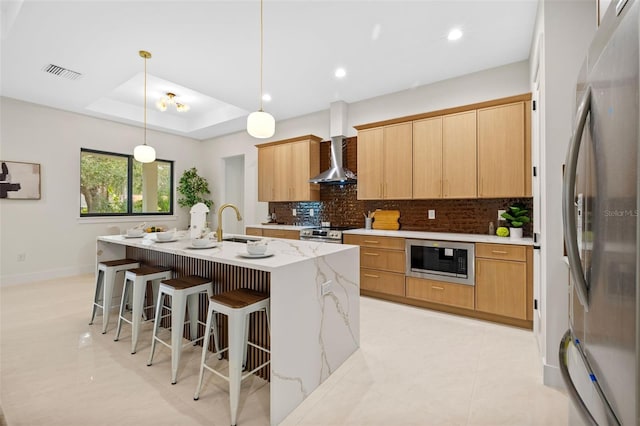 kitchen with pendant lighting, wall chimney range hood, stainless steel appliances, a kitchen island with sink, and a tray ceiling