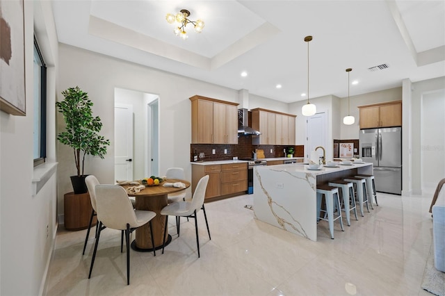 kitchen with wall chimney exhaust hood, stainless steel appliances, an island with sink, backsplash, and a tray ceiling