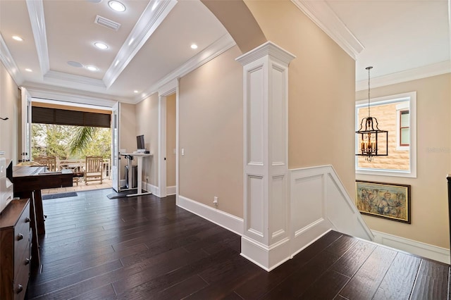 corridor featuring dark wood-type flooring, an inviting chandelier, a raised ceiling, decorative columns, and crown molding