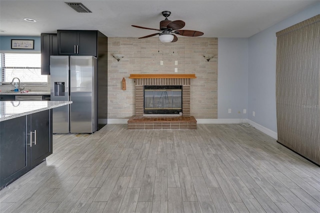 kitchen featuring ceiling fan, a fireplace, stainless steel refrigerator with ice dispenser, light wood-type flooring, and light stone counters