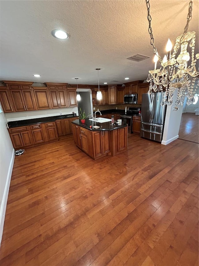 kitchen with a textured ceiling, appliances with stainless steel finishes, an island with sink, sink, and hanging light fixtures