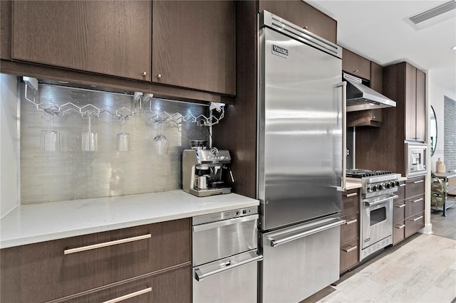 kitchen featuring tasteful backsplash, light hardwood / wood-style floors, built in appliances, light stone countertops, and dark brown cabinets