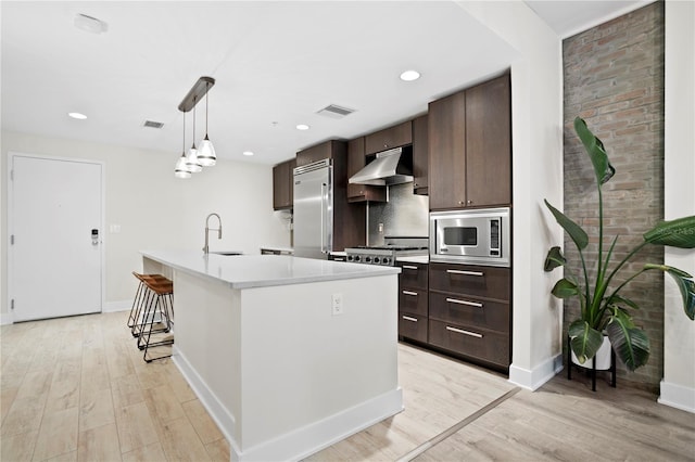 kitchen with pendant lighting, built in appliances, an island with sink, sink, and light hardwood / wood-style flooring