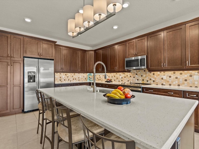 kitchen with tasteful backsplash, appliances with stainless steel finishes, an island with sink, pendant lighting, and light stone countertops