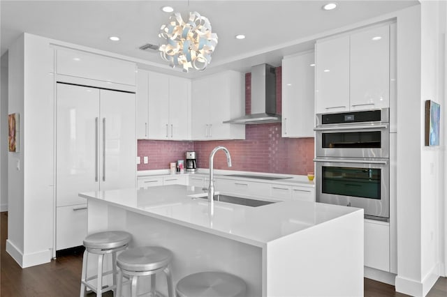 kitchen featuring white cabinetry, a center island with sink, pendant lighting, wall chimney exhaust hood, and double oven