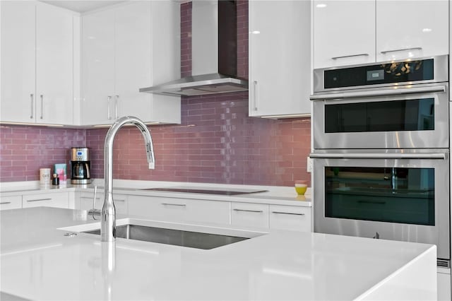 kitchen with white cabinetry, backsplash, stainless steel double oven, and wall chimney exhaust hood