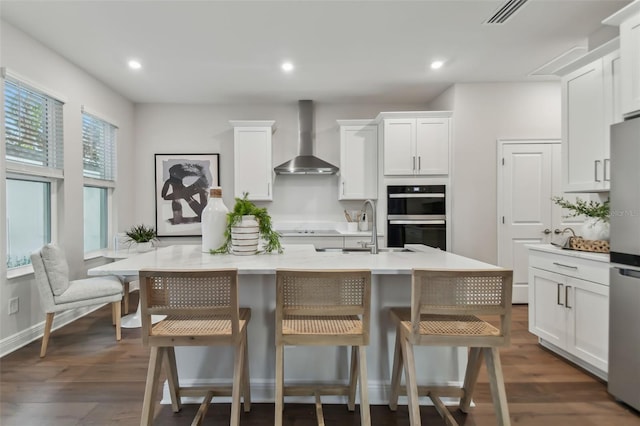 kitchen with a kitchen breakfast bar, wall chimney range hood, double wall oven, and an island with sink