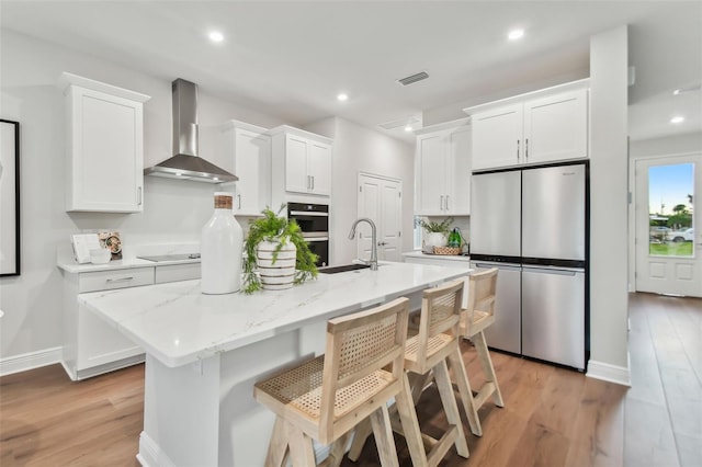 kitchen with white cabinets, wall chimney range hood, black cooktop, stainless steel fridge, and a center island with sink