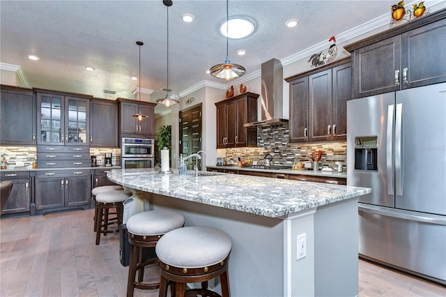 kitchen with an island with sink, wall chimney range hood, appliances with stainless steel finishes, and hanging light fixtures