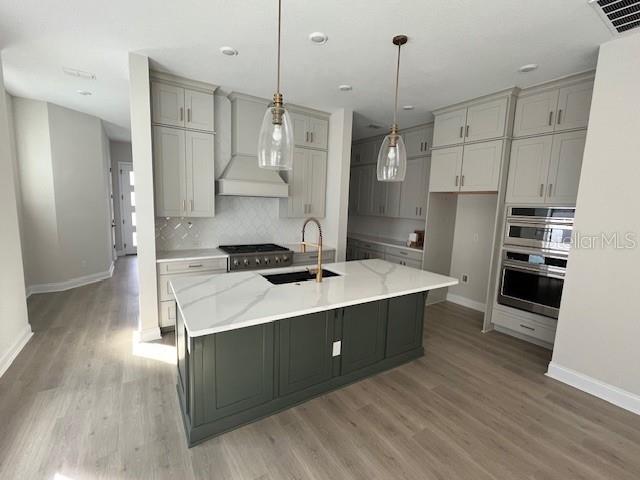 kitchen with a center island with sink, custom range hood, hanging light fixtures, a sink, and light stone countertops