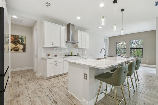 kitchen featuring white cabinets, decorative light fixtures, wall chimney range hood, sink, and a kitchen island with sink