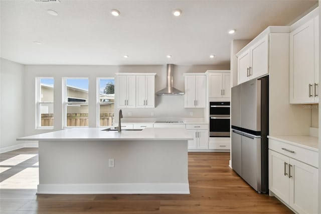 kitchen featuring an island with sink, stainless steel appliances, wall chimney range hood, white cabinets, and sink