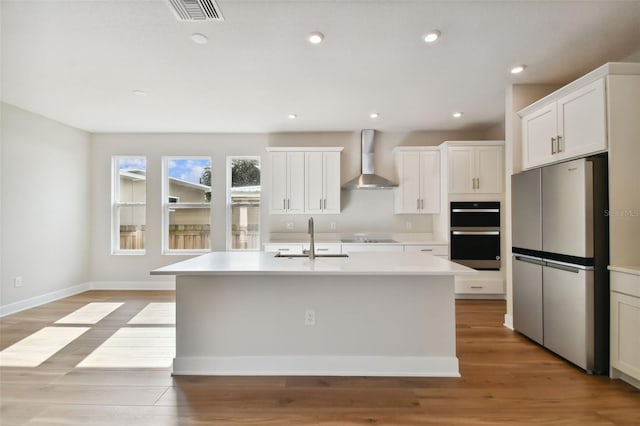 kitchen featuring white cabinets, black appliances, wall chimney exhaust hood, an island with sink, and sink