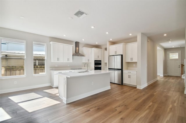 kitchen with white cabinetry, a center island with sink, stainless steel fridge, wall chimney exhaust hood, and light hardwood / wood-style flooring