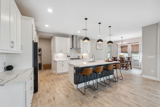 kitchen with pendant lighting, sink, white cabinets, a kitchen island with sink, and wall chimney range hood