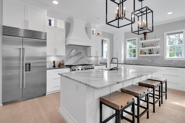 kitchen with range, white cabinetry, stainless steel built in refrigerator, sink, and custom range hood