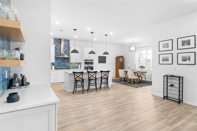 kitchen featuring backsplash, wall chimney range hood, white cabinetry, hanging light fixtures, and a breakfast bar area